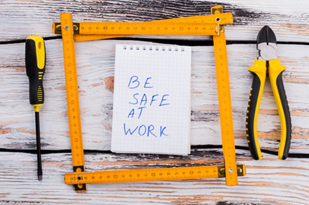 Set de unelte galben-negre, inclusiv șurubelniță, clește și metru pliabil, aranjate în jurul unui caiet cu mesajul „Be Safe at Work” scris de mână.